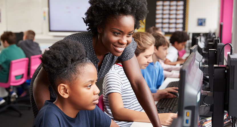 An Opus Interactive blog post image of kids working in a computer lab, there is a teacher helping the kid closest to the camera and smiling.