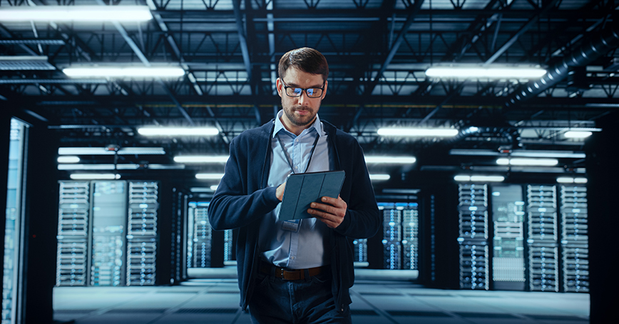 An Opus Interactive blog post image of a man working in a server room, walking towards the camera while accessing his tablet.
