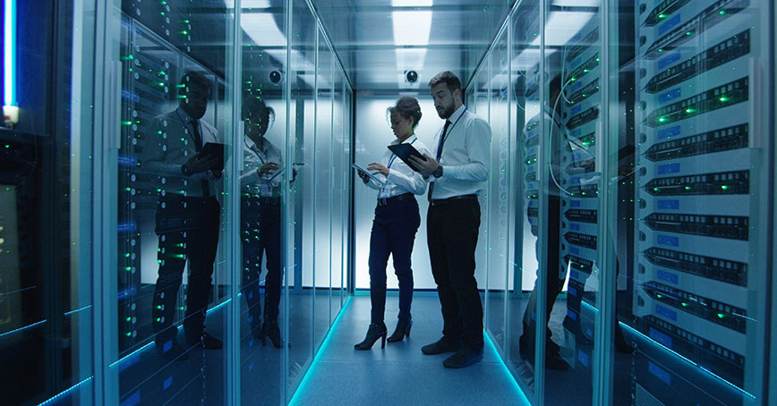 Photo of a man and woman inside a datacenter cage surrounded by cabinets and IT infrastructure, they are consorting their tablets.