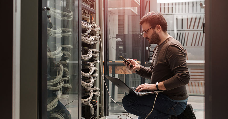 A blog post image that illustrates an IT technician kneeled in front of an open server, organizing wires as well as consorting his laptop.
